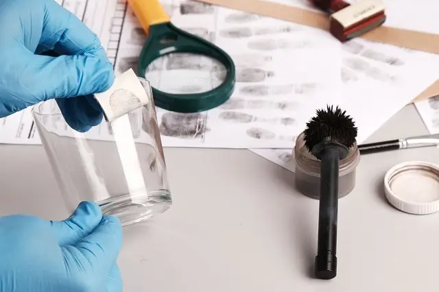 A forensic investigator lifts a fingerprint from a glass using tape, with fingerprint samples and tools on the table.