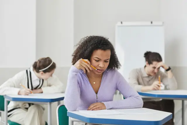 a young girl with curly hair, holds a pencil to her mouth and appears to be deep in thought as she looks down at her exam paper