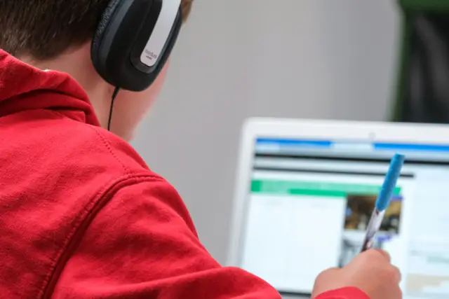 A young person in a red hoodie is sitting in front of a laptop, wearing black over-the-ear headphones