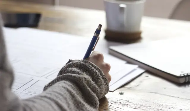 person writing on brown wooden table near white ceramic mug
